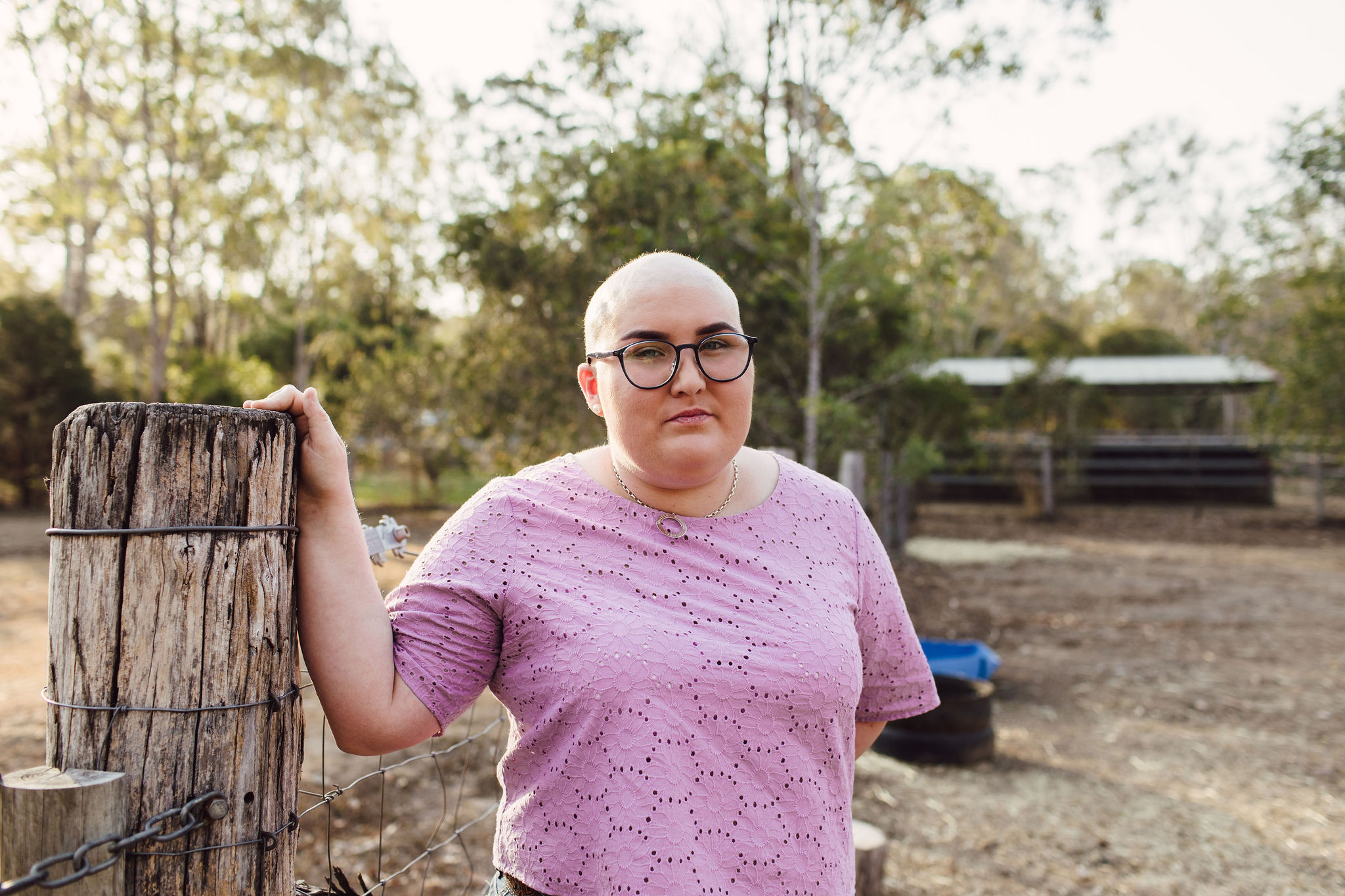 Woman with a shaved head stands outside in a large, leafy yard