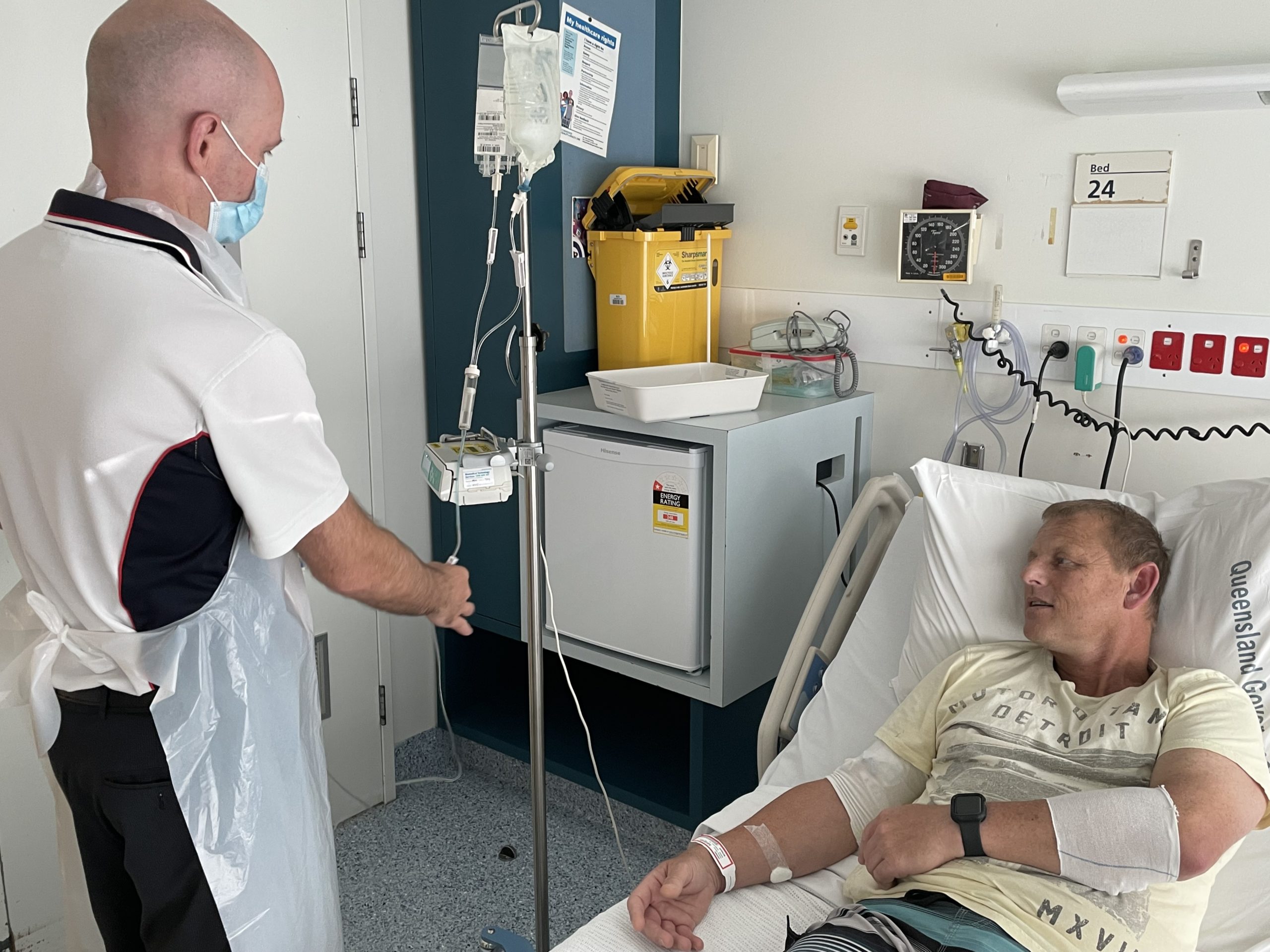 Graeme lies in a hospital bed while a nurse connects an IV line to him