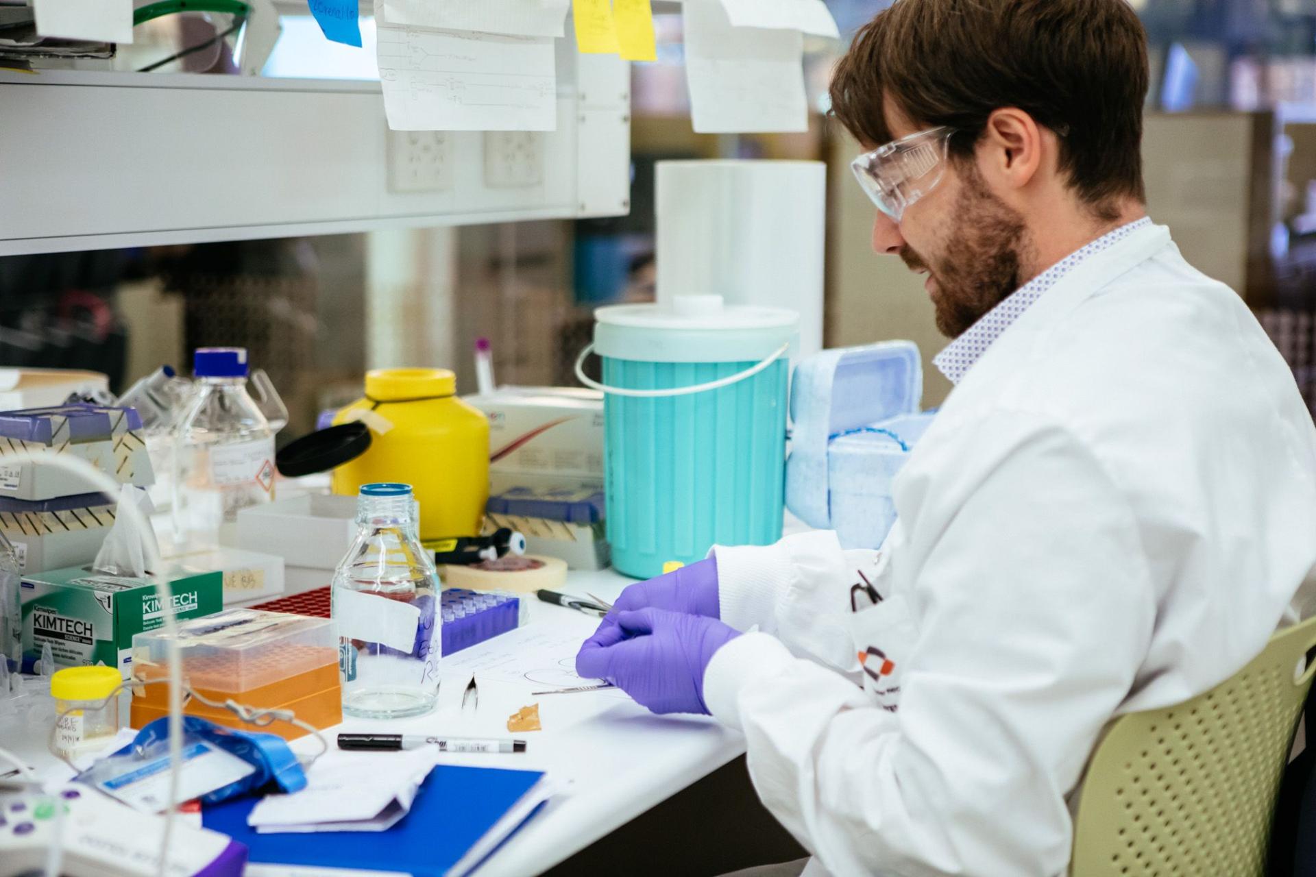 A male scientist in a white lab coat, safety goggles, and purple gloves works with tweezers at a cluttered lab bench filled with various scientific equipment.